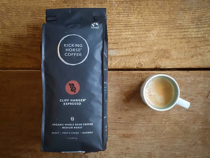 Bag of Kicking Horse Coffee, Cliff Hanger Espresso roast lying flat on table beside cup of coffee
