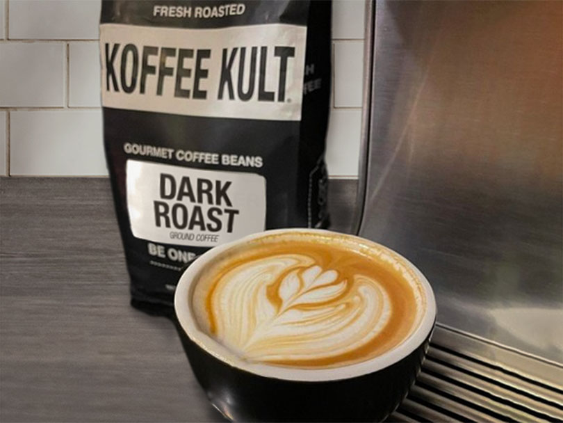 Koffee Kult dark roast gourmet coffee beans to the side of espresso machine with cup of freshly made cappuccino