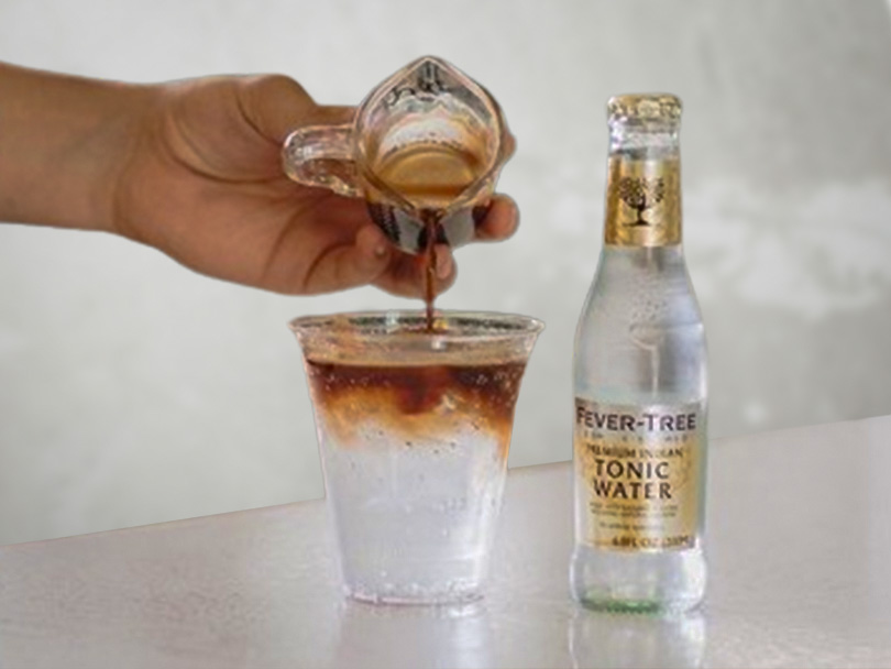 Hand pouring espresso into Fever-Tree tonic water and ice, demonstrating how to make espresso tonic