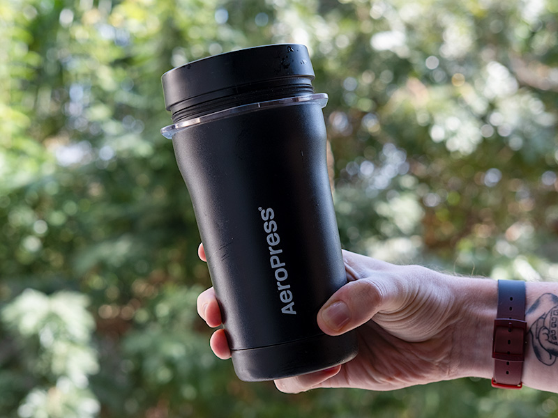 Man's hand holding the fully packed AeroPress Go Plus insulated mug out in front of a green bush