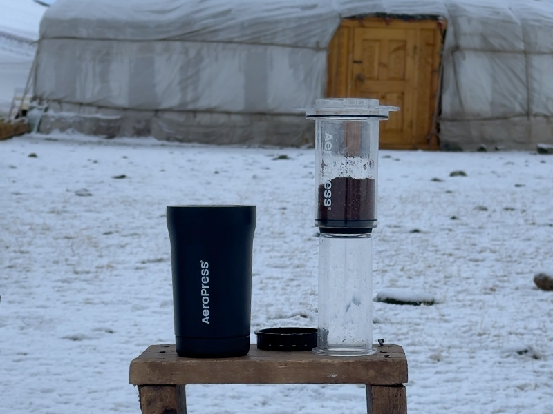 AeroPress brewer filled with ground coffee, thermal Go Plus mug to the side. Set in a snowy field with a Mongolian yurt in the background