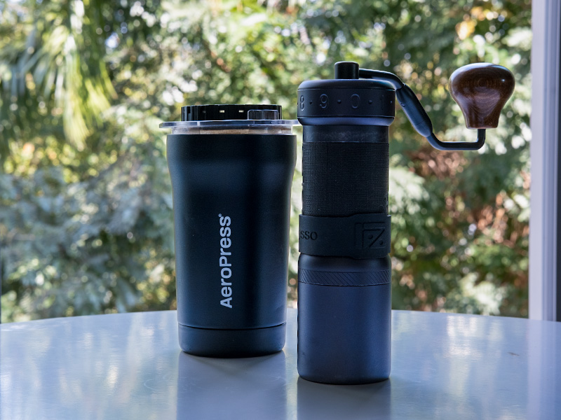 AeroPress Go Plus and 1Zpresso K Ultra sitting side-by-side on table with green bushes outside the window