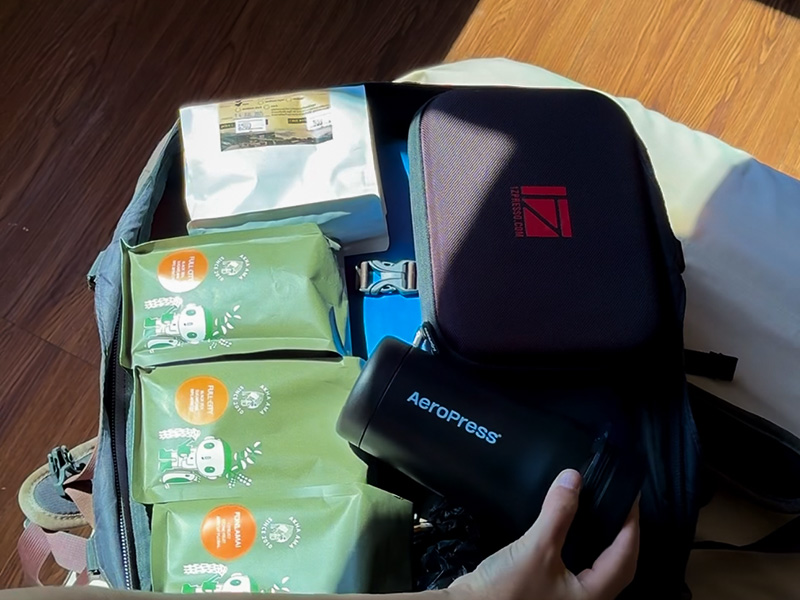 Hand placing the AeroPress Go Plus into a 40L backpack alongside 4 bags of coffee and an 1Zpresso grinder carry case