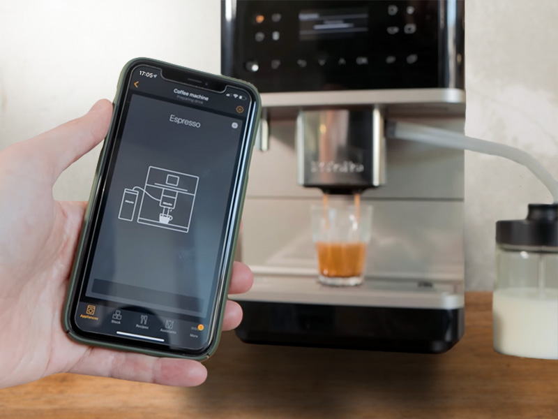 Hand holding an iPhone with Miele App open in front of the CM 6360 which is making an espresso, ordered from the app