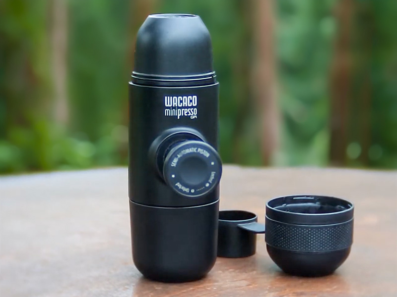 Wacaco Minipresso GR sitting on wooden bench at forest campsite, lid and scoop beside the espresso maker