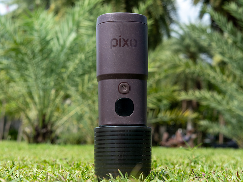 Wacaco Pixapresso standing upright with attached cup, in the grass with trees behind