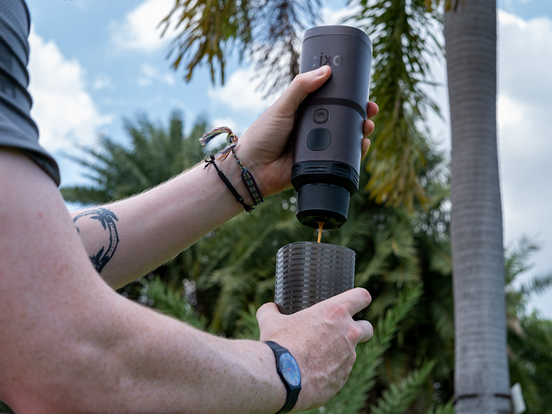 Wacaco Pixapresso mid-way through making an espresso shot, mans hands holding it in the park, with trees and blue sky in background