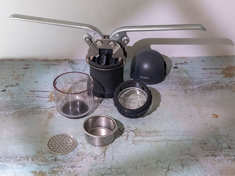 All the parts for the HUGH Leverpresso Lite V4 laid out on a table (lever espresso maker, lid, cup, shower screen, unpressurized filter basket, basket holder)