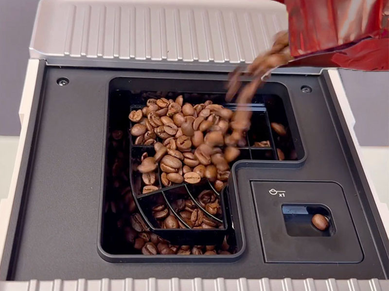 Pouring medium-dark whole coffee beans from a bag into the top-loading bean hopper of the KitchenAid KF3, a stray bean is sitting on the pre-ground coffee chute's lid