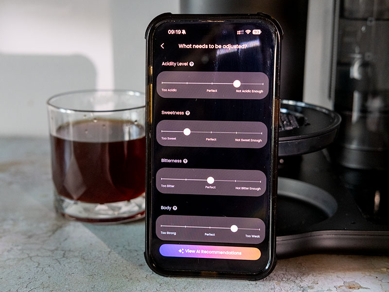 Phone with Cosori Juni's VeSync app opened, displayed a screen asking 'What needs to be adjusted?' and sliders for Acidity Level, Sweetness, Bitterness, and Body. The button at the bottom says 'View AI Recommendations' to improve future cups of pour over coffee