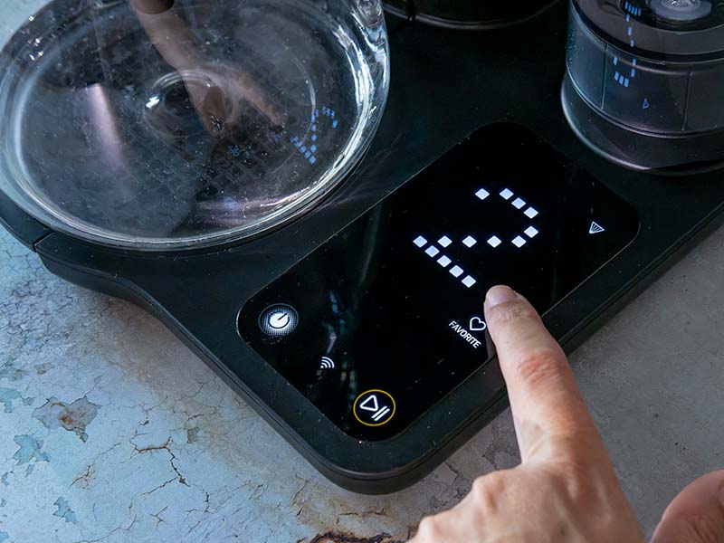 Finger pressing the 'Favorite' button on the Cosori Juni's touchscreen with a big '2' displayed on the screen to indicate which recipe has been selected