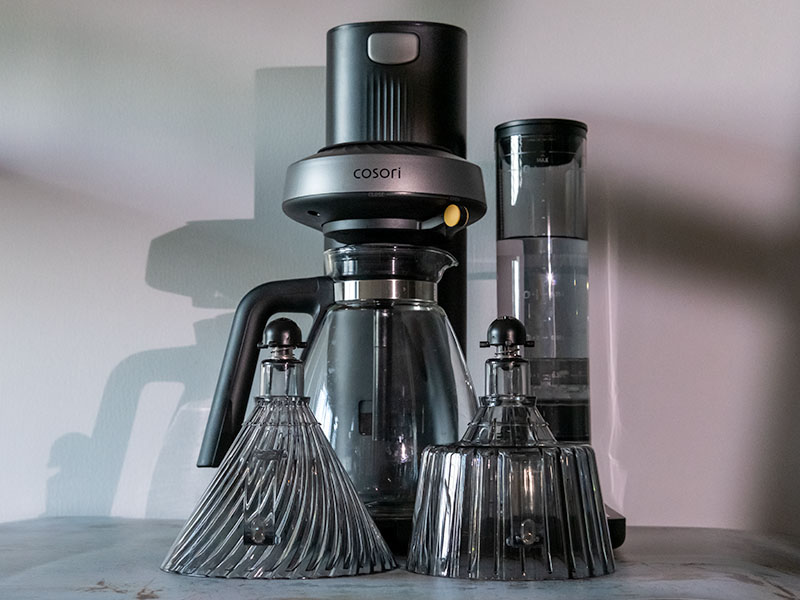 Straight on view of the Cosori Juni, with 2 coffee filter baskets (cone-shaped one for 1-2 cups, and flat-bottomed one for the carafe) sitting in front of it