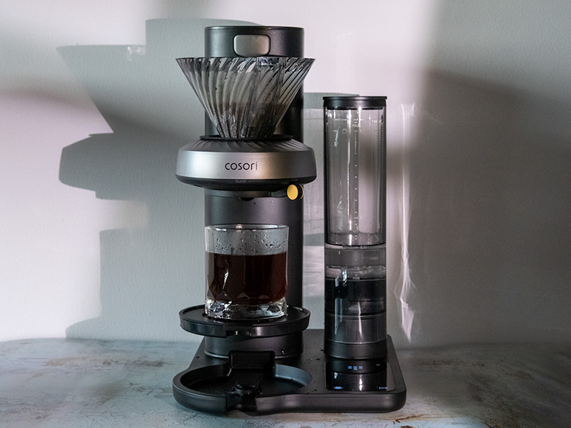 Front view of the Cosori Juni automatic pour over machine, with a fresh glass of coffee sitting on the drip tray
