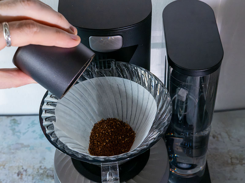 Pouring ground coffee from grinder catch cup into the filter for the Cosori Juni ahead of brewing
