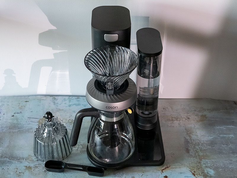 Top-down view of the Cosori Juni, set up for auto pour over, spare basket and coffee scoop sitting beside the machine