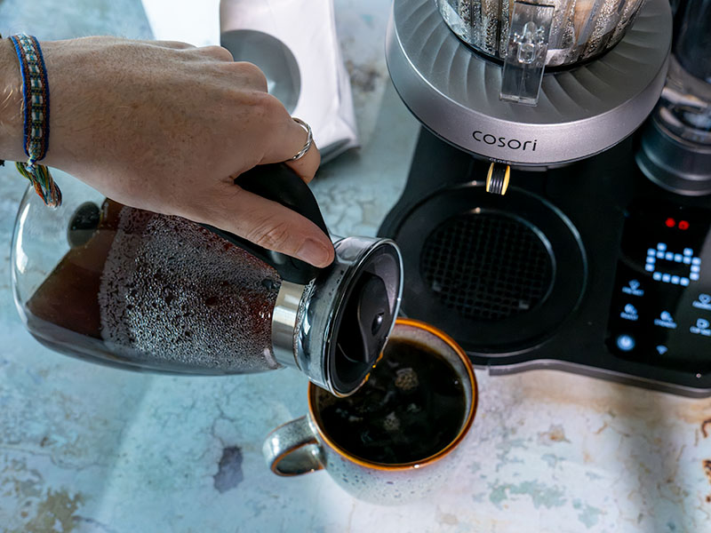 Hand pouring pour over coffee from carafe into a mug with the Cosori Juni machine turned on in the background