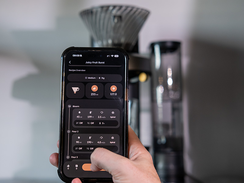 Hand holding phone in front of the Cosori Juni, the VeSync app is open on 'Juicy Fruit Burst' coffee recipe